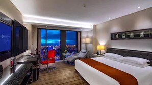 Une chambre d’hôtel avec un lit, un bureau, une télévision, une chaise et une vue sur la ville la nuit.