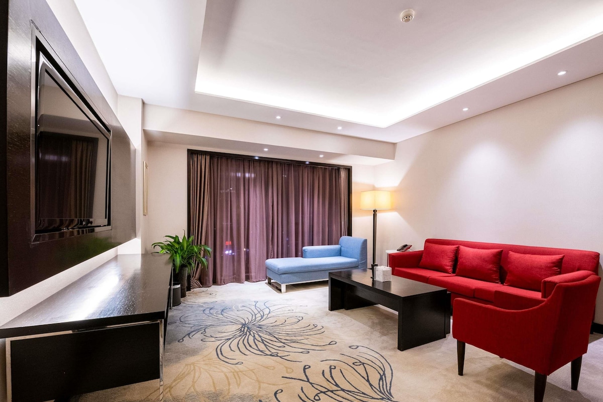 Quarto de hotel moderno com televisão de tela plana, um sofá vermelho, um sofá azul, uma mesa de centro, uma luminária e um tapete decorativo.