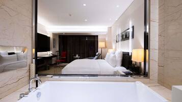 A modern hotel room with a large bathtub, a desk with a chair, a red chair, and a bed with white bedding.