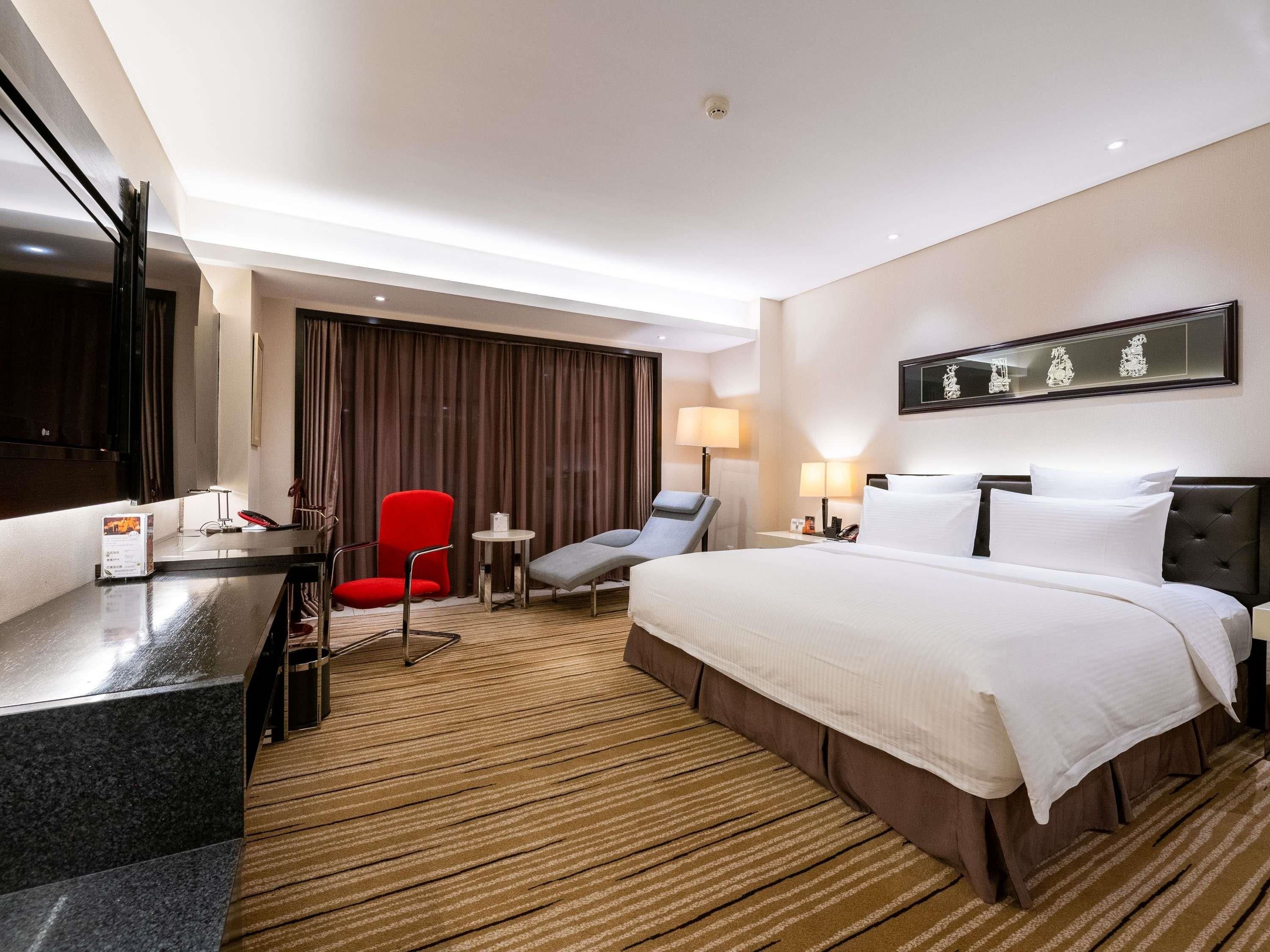 Una camera d'hotel con un letto, una scrivania, una poltrona rossa, una poltrona grigia, un tavolino e una lampada a parete.