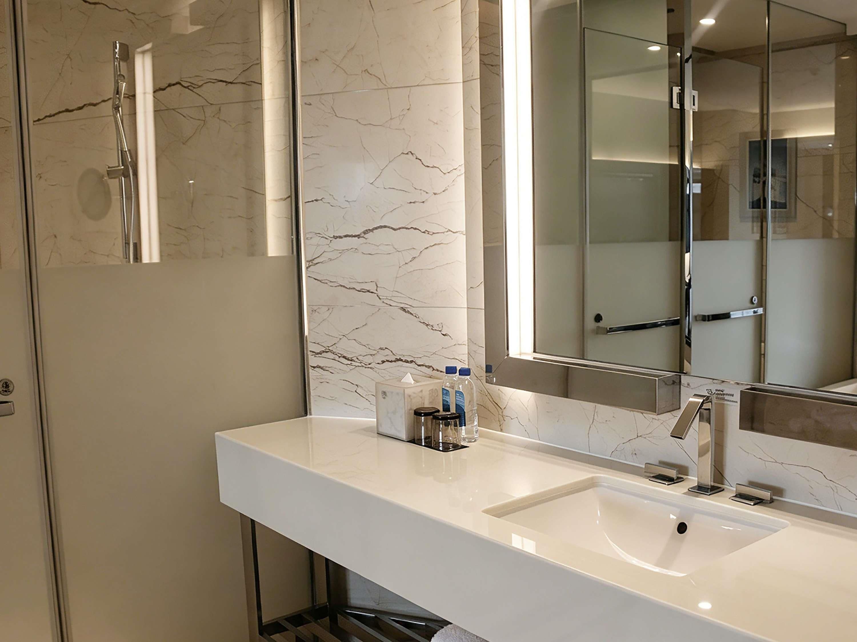 A modern bathroom with a marble wall, a large mirror, a shower, and a white sink with toiletries.