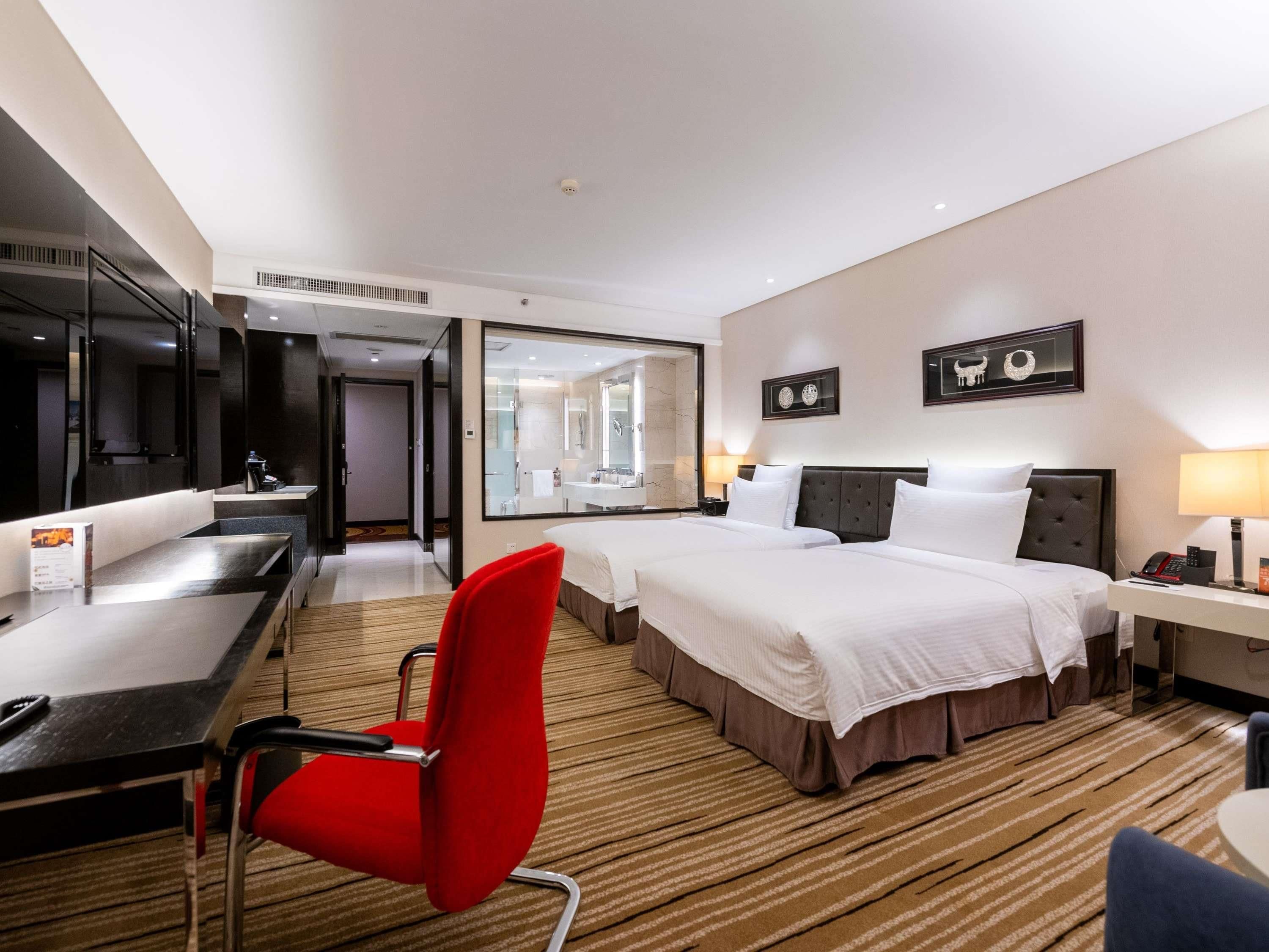 A hotel room with a bed, a desk, a chair, a TV, and a bathroom visible through a glass door.