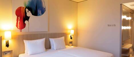 A hotel room with a bed, two bedside lamps, a wall-mounted artwork, and a mirror with lights.