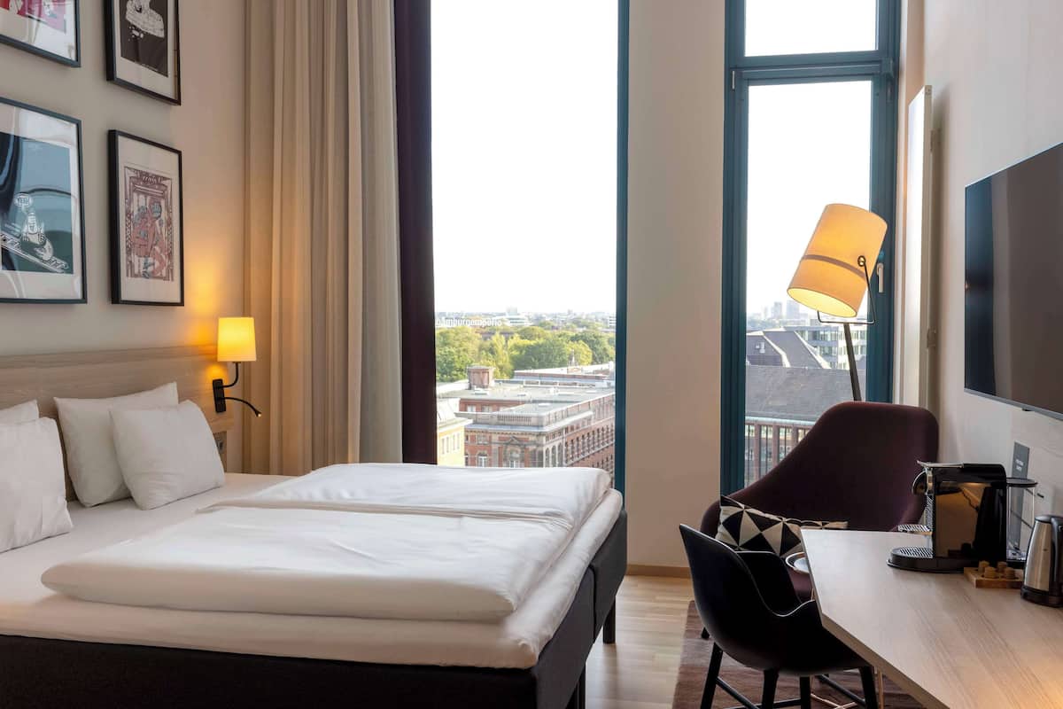 A hotel room with a bed, a desk with a chair, a lamp, and a TV.