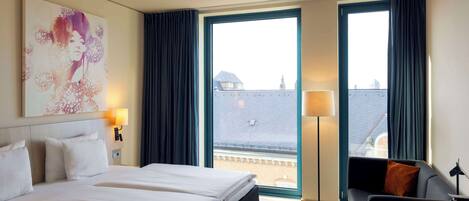 Habitación de hotel con una cama grande, dos mesitas de noche, un escritorio y una silla. Cuenta con un ventanal con cortinas, una lámpara de pie y un cuadro en la pared.