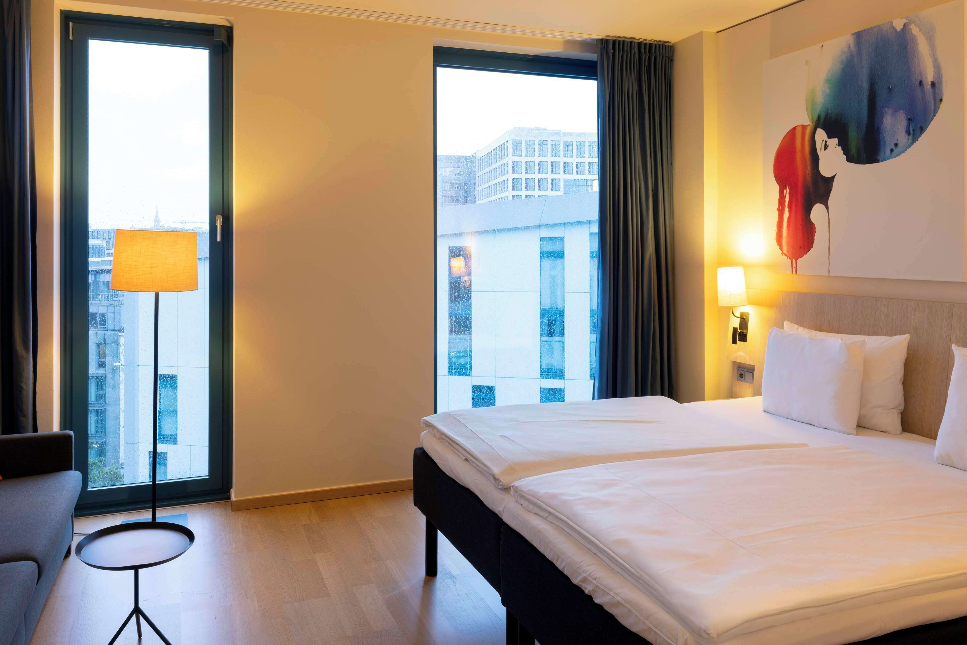 Ein Hotelzimmer mit einem großen Bett, einem kleinen Tisch, einer Lampe und Blick auf die Stadt durch die Fenster.