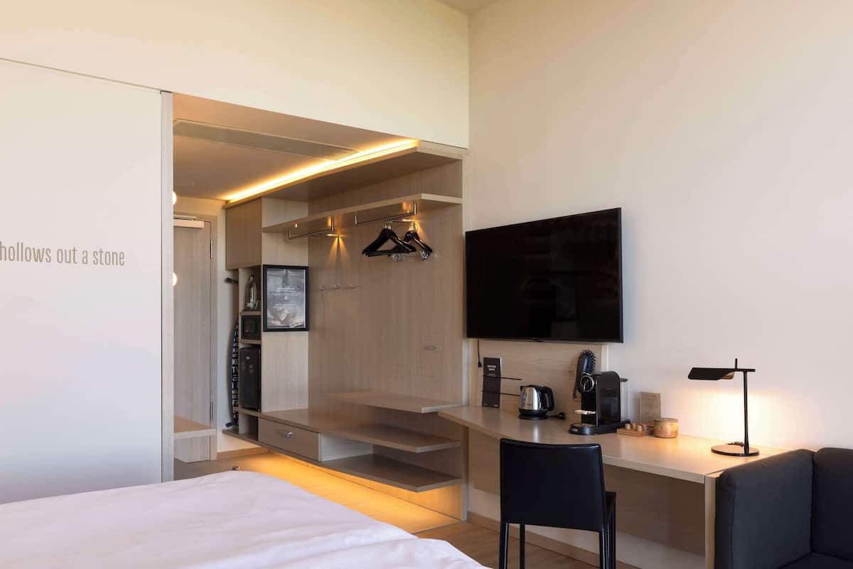 A modern hotel room with a bed, a desk, a chair, a TV, and a kitchenette area.