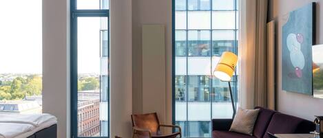 Una habitación de hotel moderna con un ventanal grande, una cama, un sofá, una mesita y una lámpara.