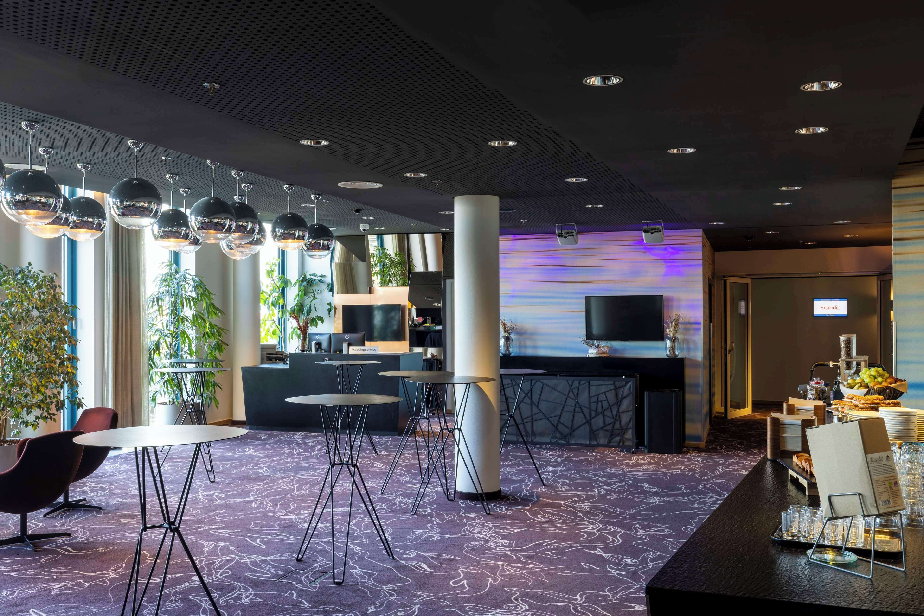 A modern hotel lobby with round tables, a reception desk, and a large screen displaying a scenic view.