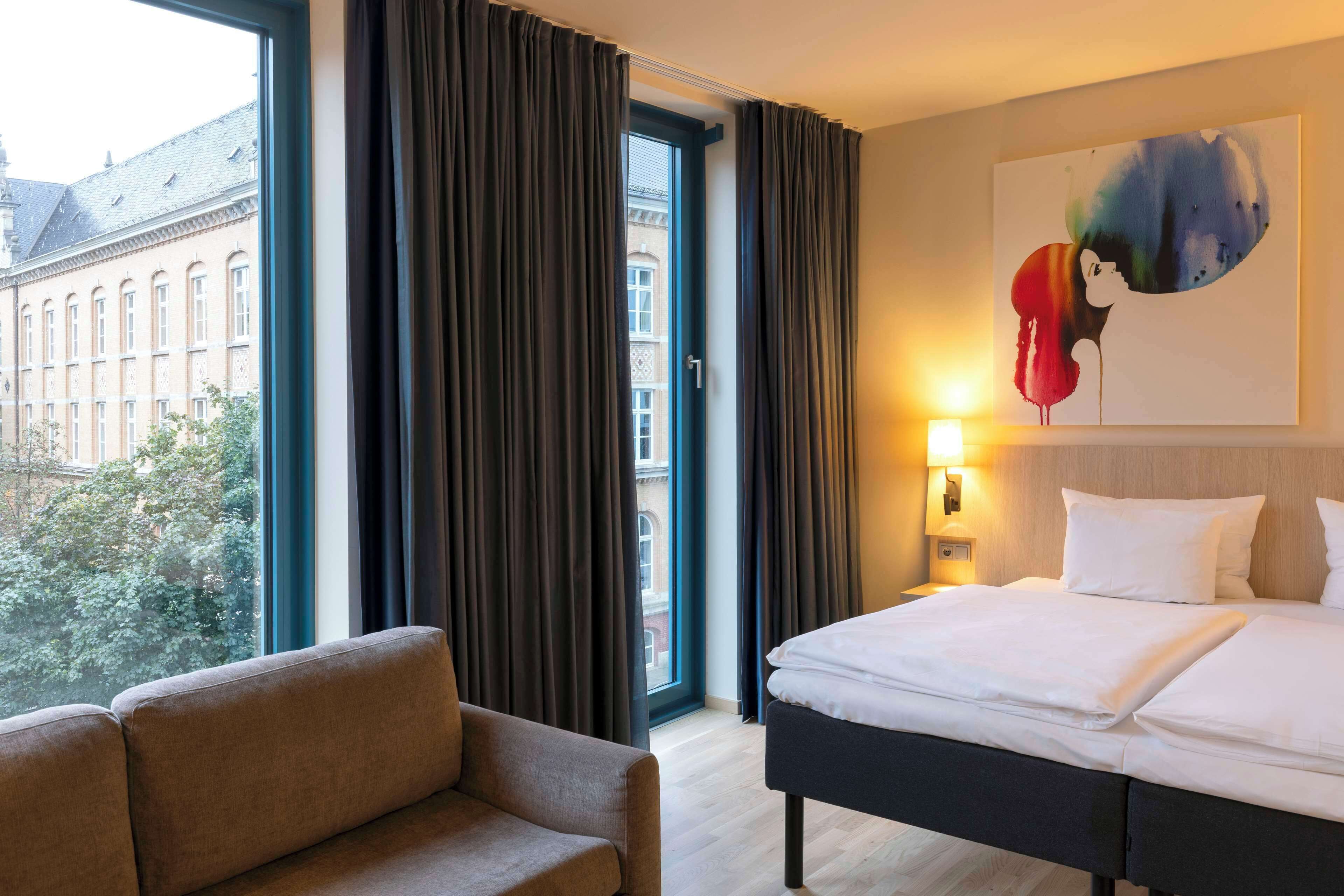 A hotel room with a bed, a sofa, a painting on the wall, and a window with curtains.