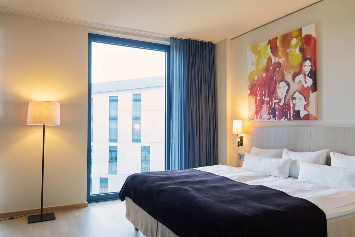 A hotel room with a large bed, a floor lamp, a painting on the wall, and a view of a building through the window.
