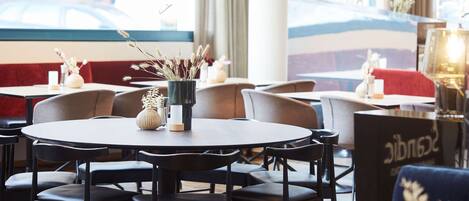 Un restaurante con mesas redondas, sillas negras y vistas a un edificio a través de amplios ventanales.