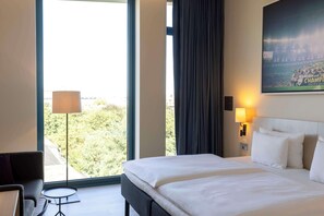 Habitación de hotel con una cama grande, un escritorio, una lámpara y vistas a árboles a través de una ventana.