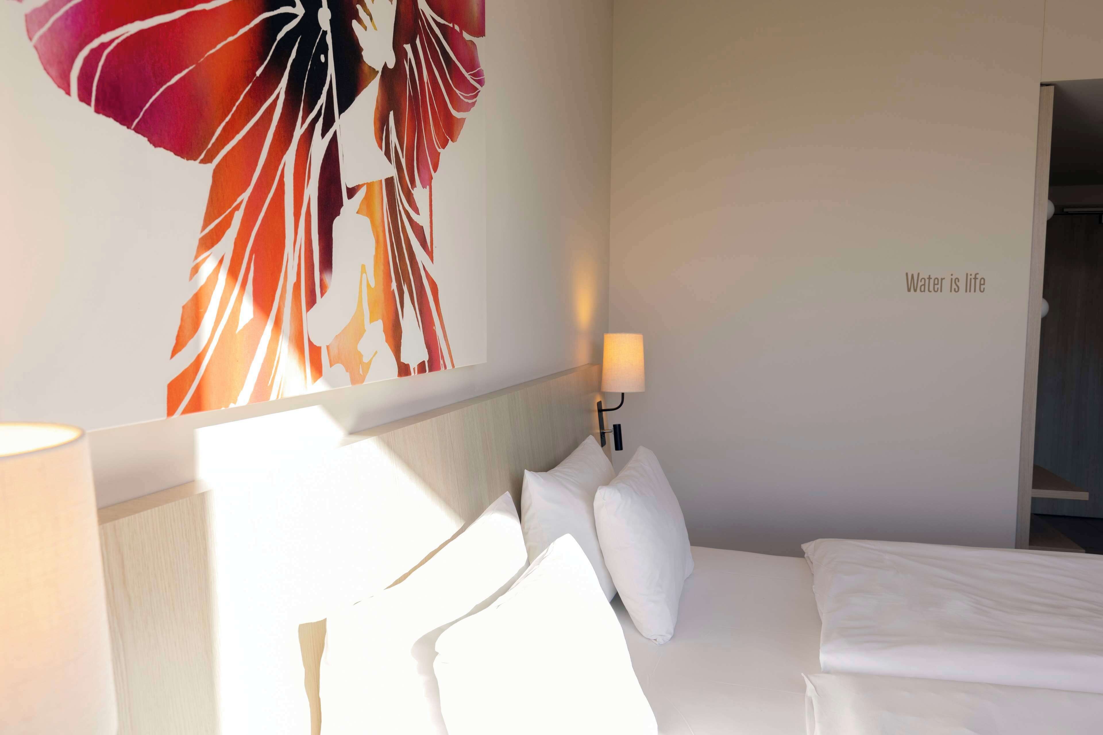 Habitación de hotel con cama, almohadas, una lámpara de pared y un cuadro abstracto grande.