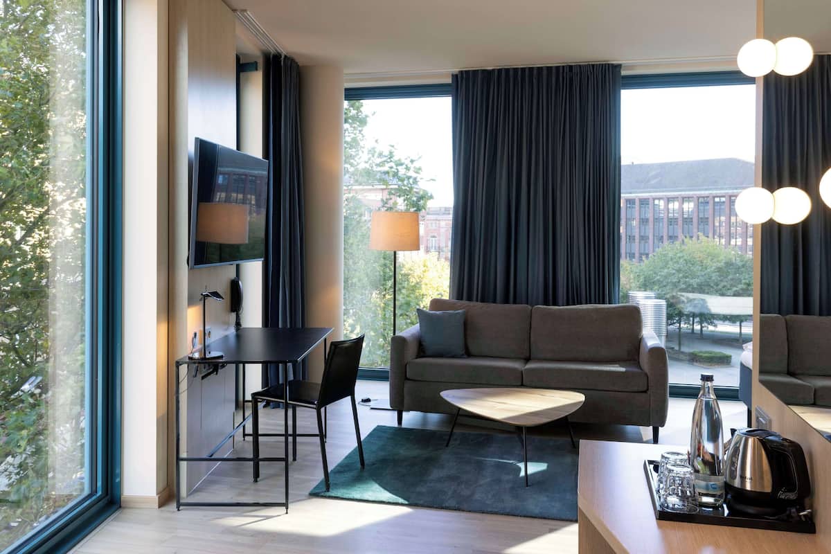 A modern hotel room with a sofa, a coffee table, a TV, and a dining area with a table and chairs.
