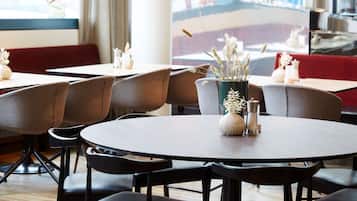 Un restaurante con mesas redondas y sillas negras, un florero con plantas secas y un amplio ventanal con vista a un edificio.