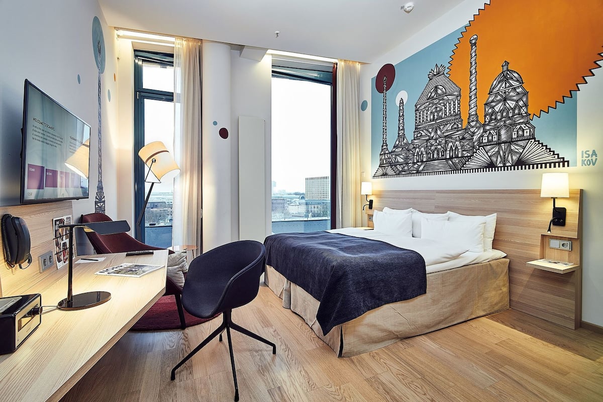 A hotel room with a bed, desk, chair, and a large window with a city view.