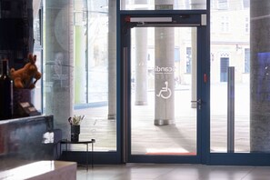 Entrada de hotel con puerta de vidrio, señal de accesibilidad para sillas de ruedas y vista de la calle.