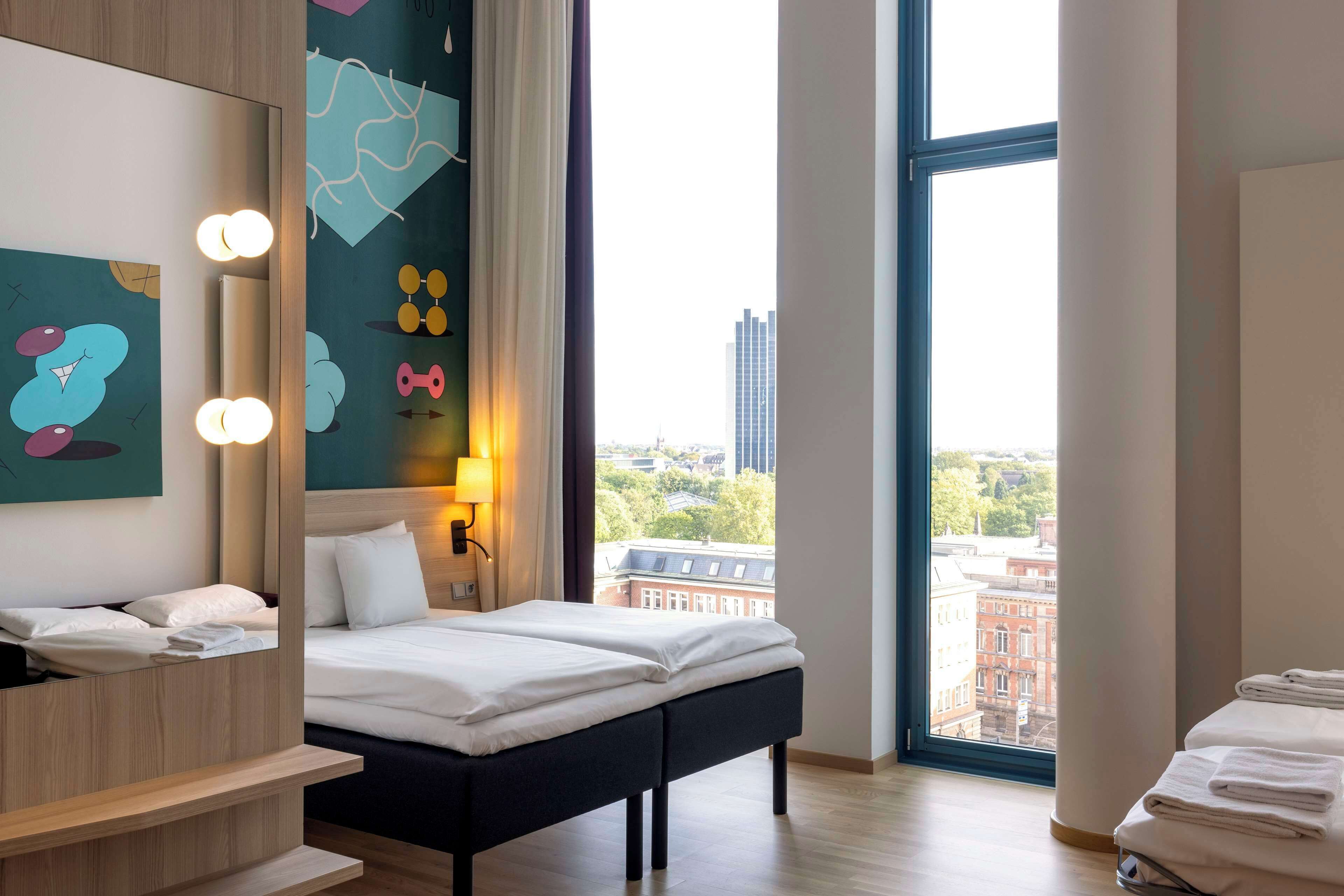 Et moderne hotelværelse med seng, natbord og stort vindue med udsigt over byen.