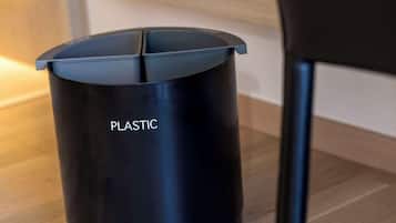 Uma lixeira preta identificada com a etiqueta 'PLASTIC' em um quarto com piso de madeira e uma cadeira preta.
