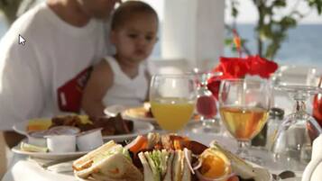 Una mesa de comedor con comida y bebidas, un niño, y vista al mar.
