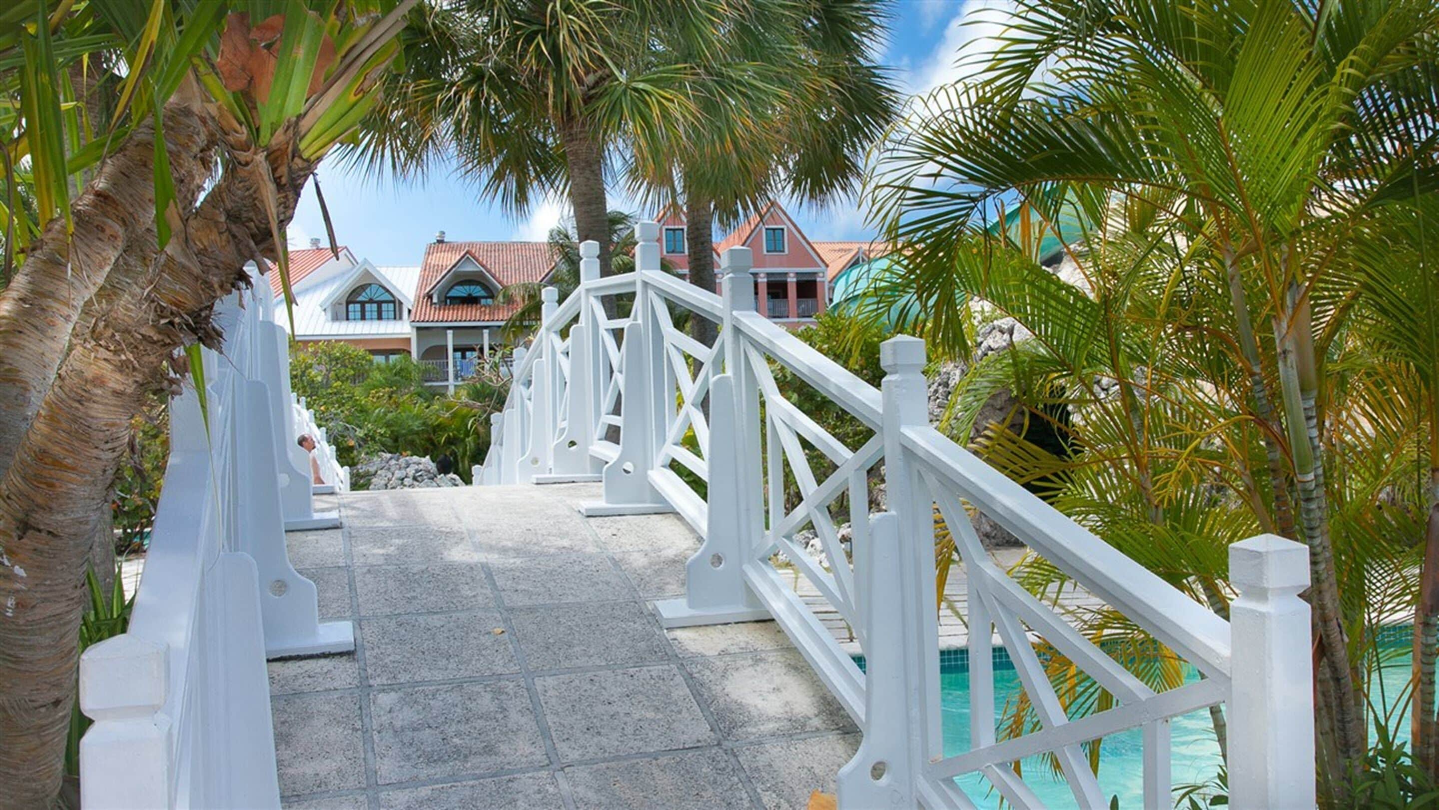 Un puente blanco que conduce a una zona de piscina con palmeras y casas al fondo.