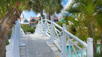 Un puente blanco que conduce a una zona de piscina con palmeras y casas al fondo.