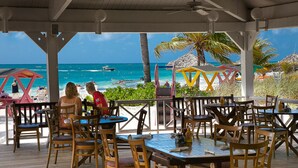 A beachside restaurant with outdoor seating overlooking the ocean, featuring wooden tables and chairs, and a view of the beach and boats. - The Marlin at Taino Beach Resort & Clubs (Freeport)