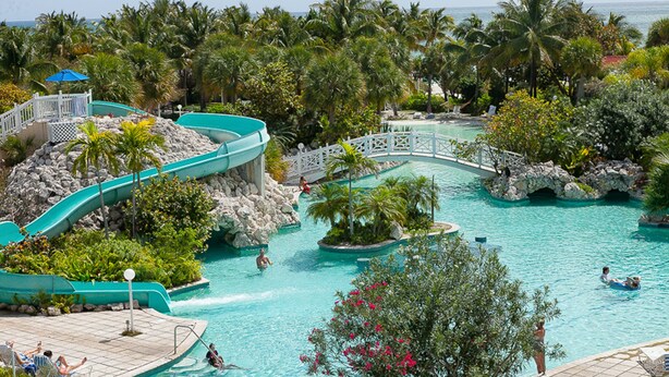A large outdoor pool with a water slide, surrounded by palm trees and rocky formations. - The Marlin at Taino Beach Resort & Clubs (Freeport)