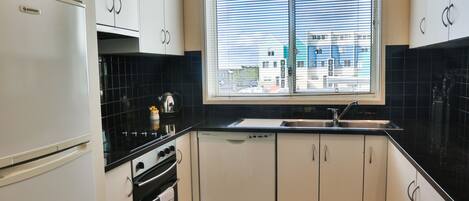 A kitchen with white cabinets, a black countertop, a window with blinds, and a view of buildings outside.