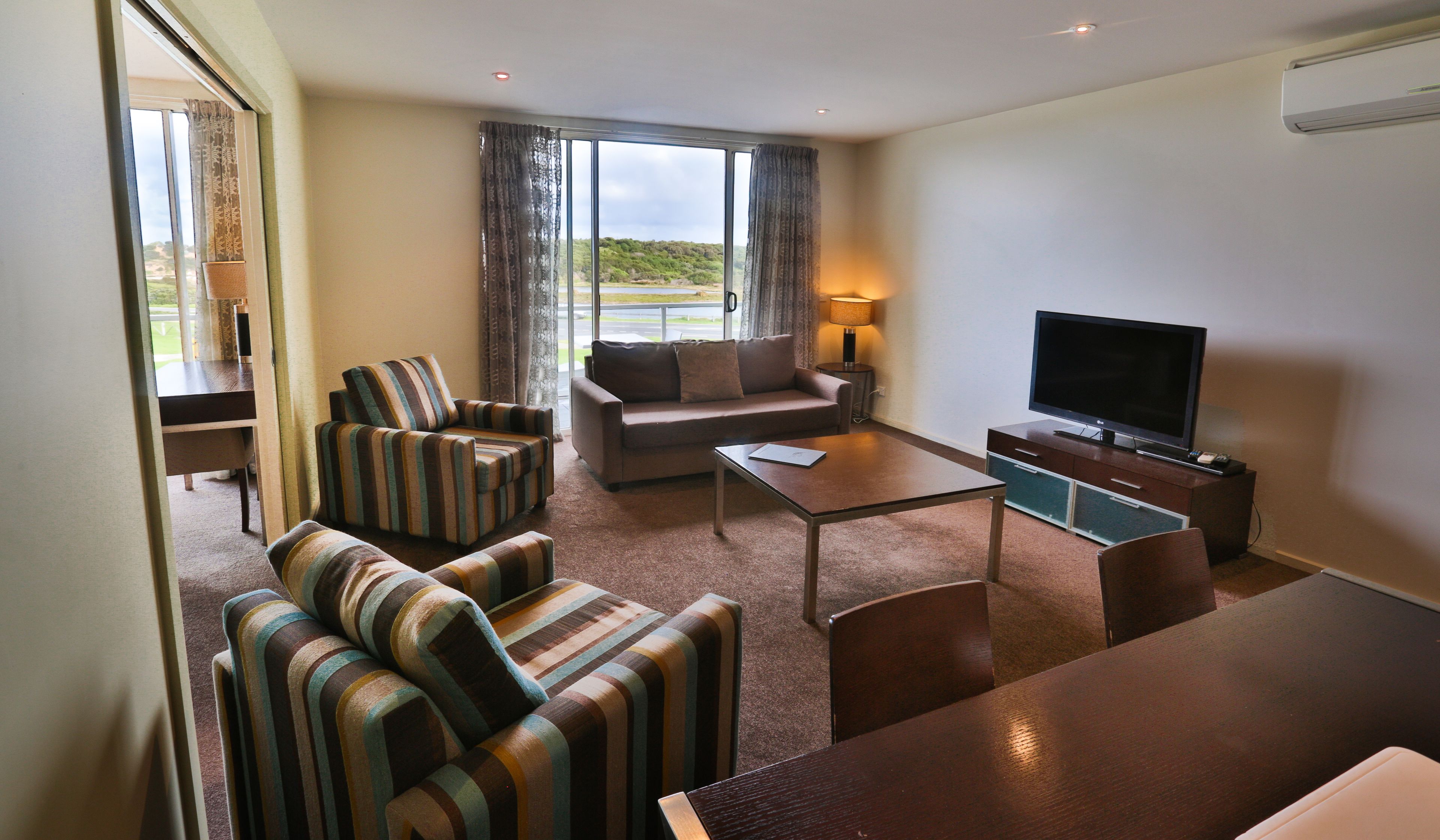 A hotel room with a sofa, armchairs, a coffee table, a TV, and a view of the outdoors.