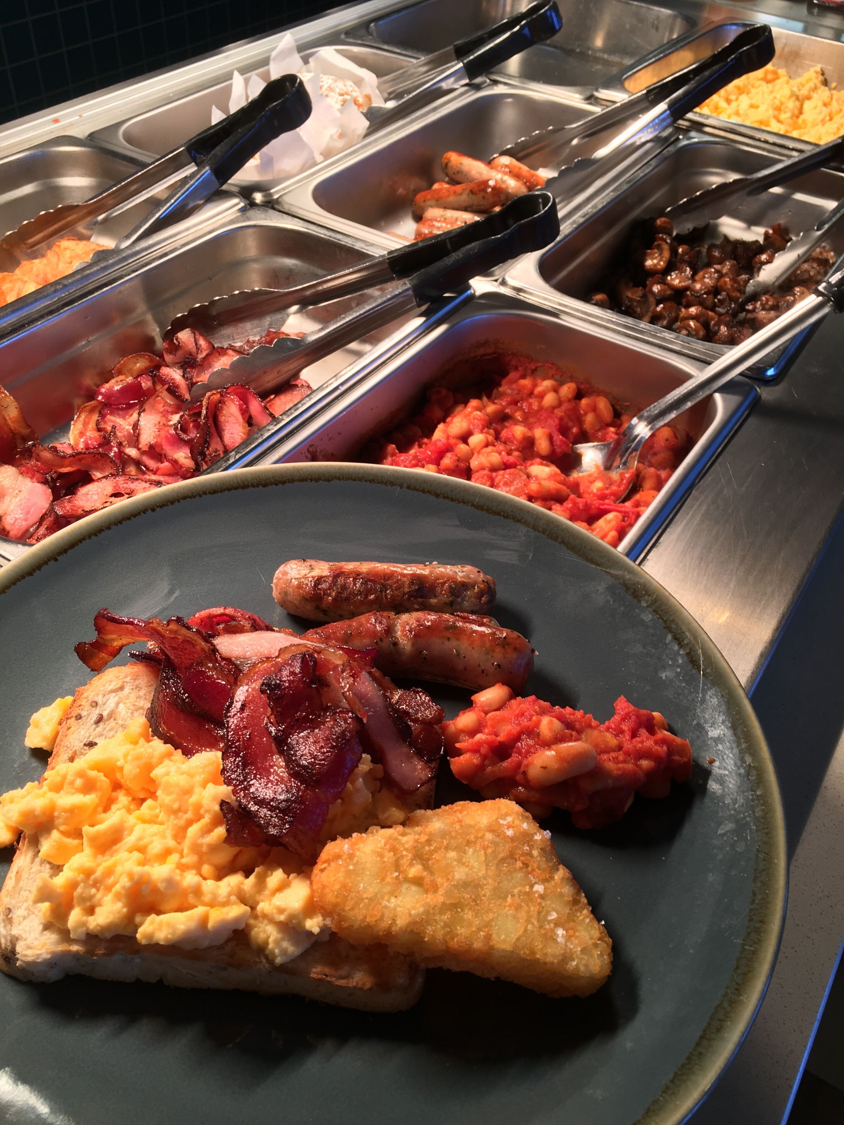 A buffet with various breakfast items including scrambled eggs, bacon, sausages, and beans.
