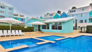Una piscina con un pequeño edificio, sillas de descanso blancas y sombrillas.
