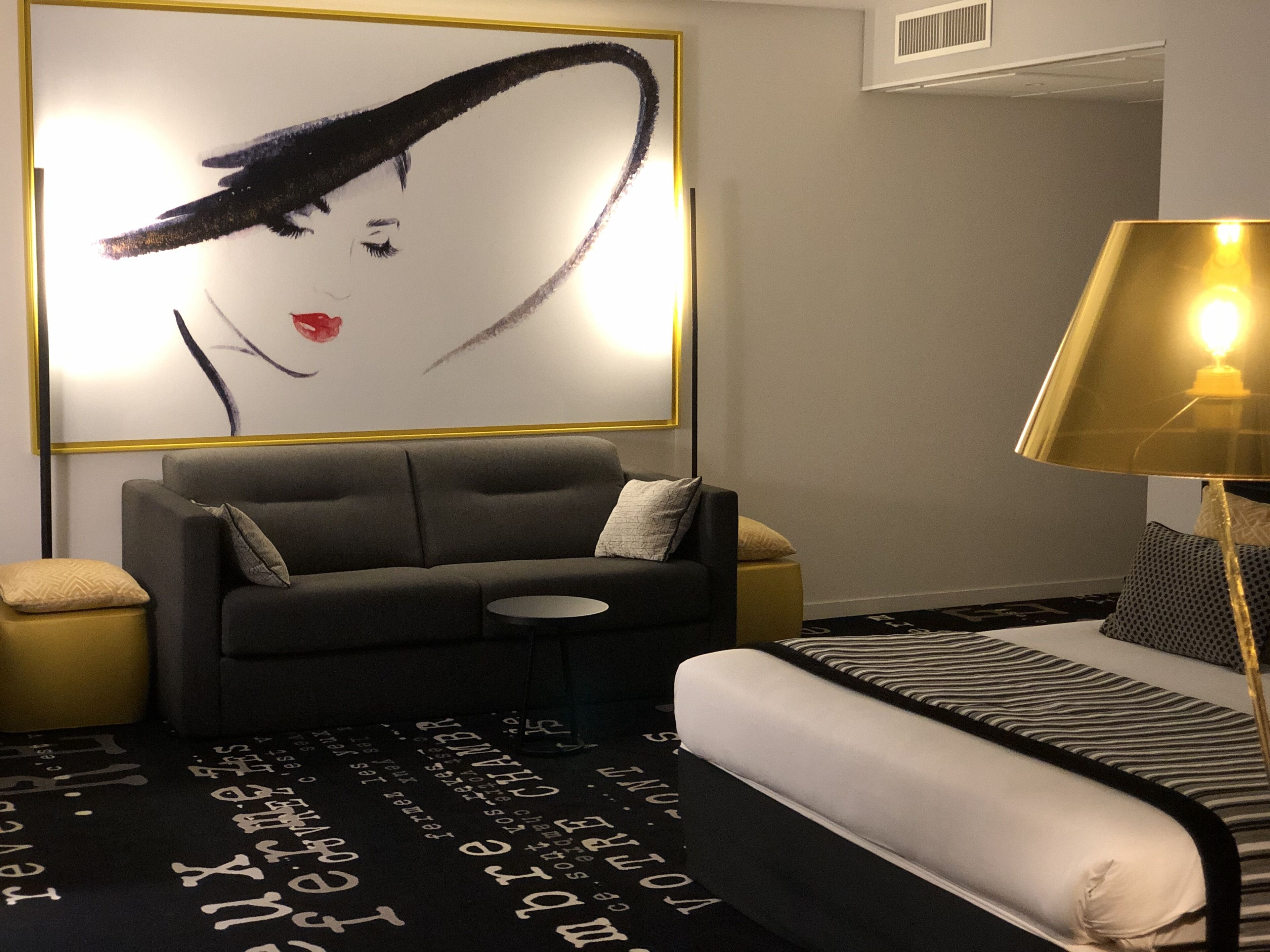 A modern hotel room with a grey sofa, a small round table, a bed with striped bedding, and a large abstract painting on the wall.