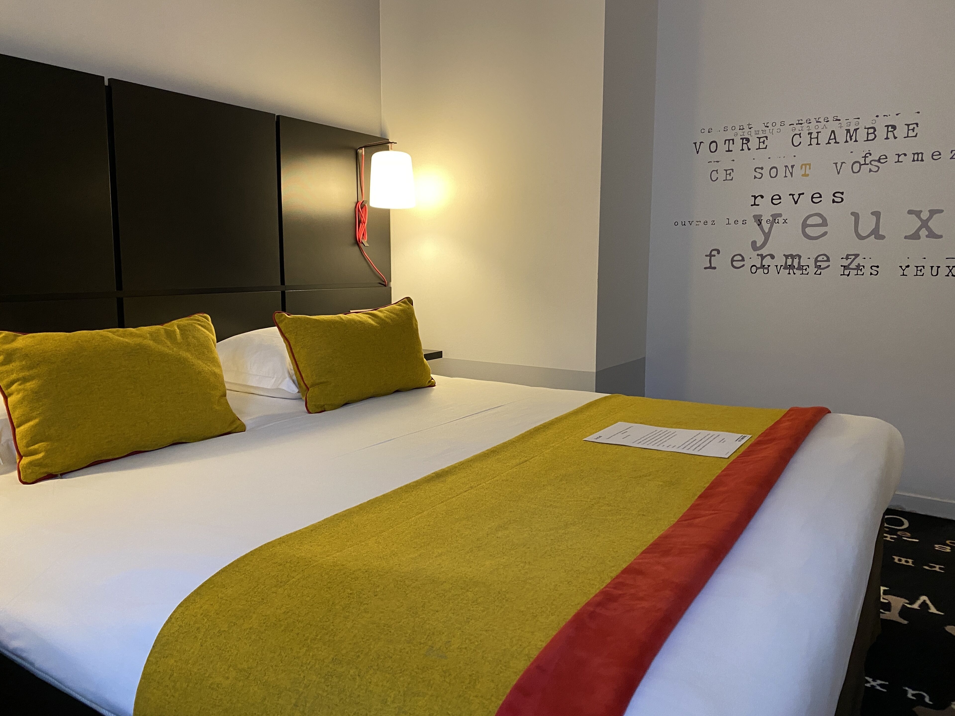 A hotel room with a neatly made bed, a headboard, a wall with text, and a lamp.