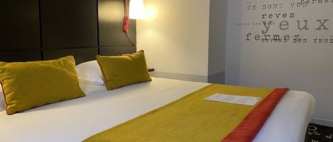 A hotel room with a neatly made bed, a headboard, a wall with text, and a lamp.