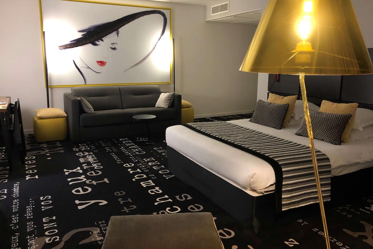 Una habitación de hotel moderna con una cama grande, un sofá, una pequeña mesa redonda y una lámpara de pie alta.