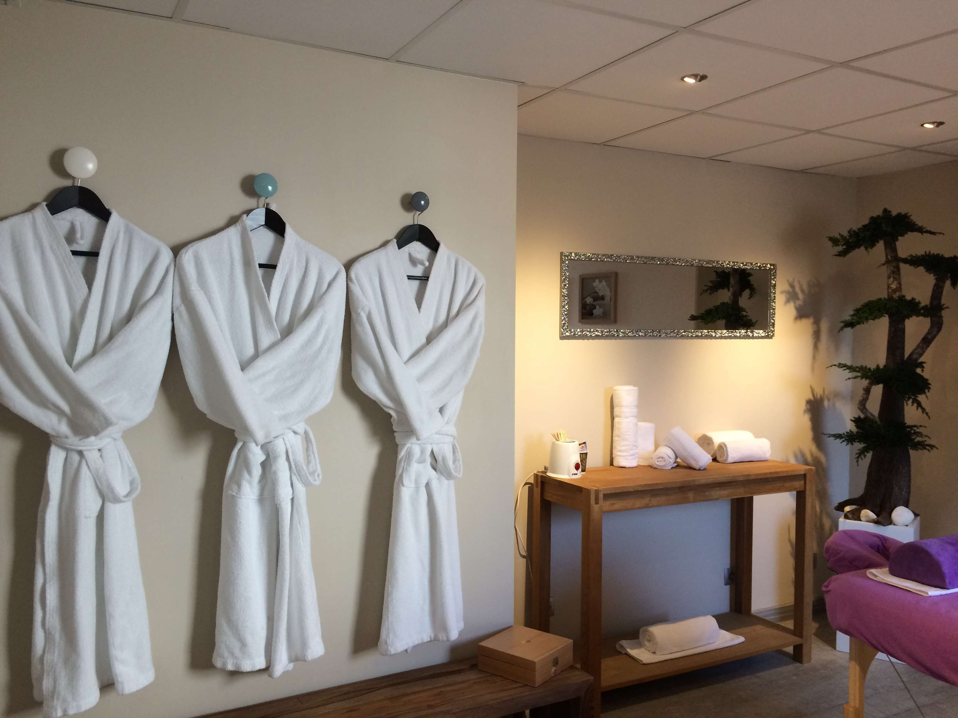 A spa room with three white robes hanging on a wall, a wooden table with towels and toiletries, and a mirror with a decorative frame.