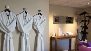 A spa room with three white robes hanging on a wall, a wooden table with towels and toiletries, and a mirror with a decorative frame.