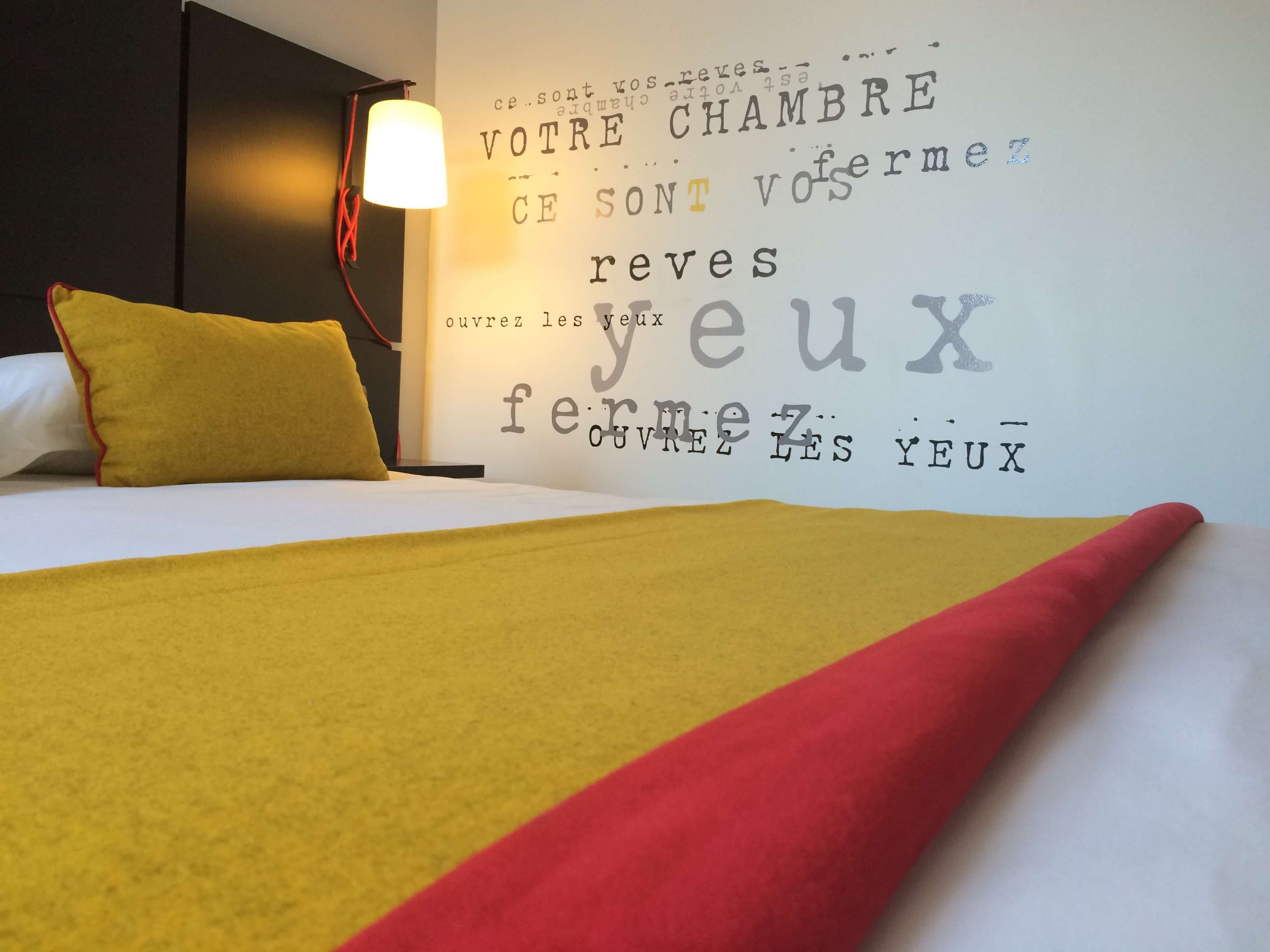 Une chambre d’hôtel avec un lit, une couverture jaune et rouge, un oreiller et un mur orné d’une phrase en français.