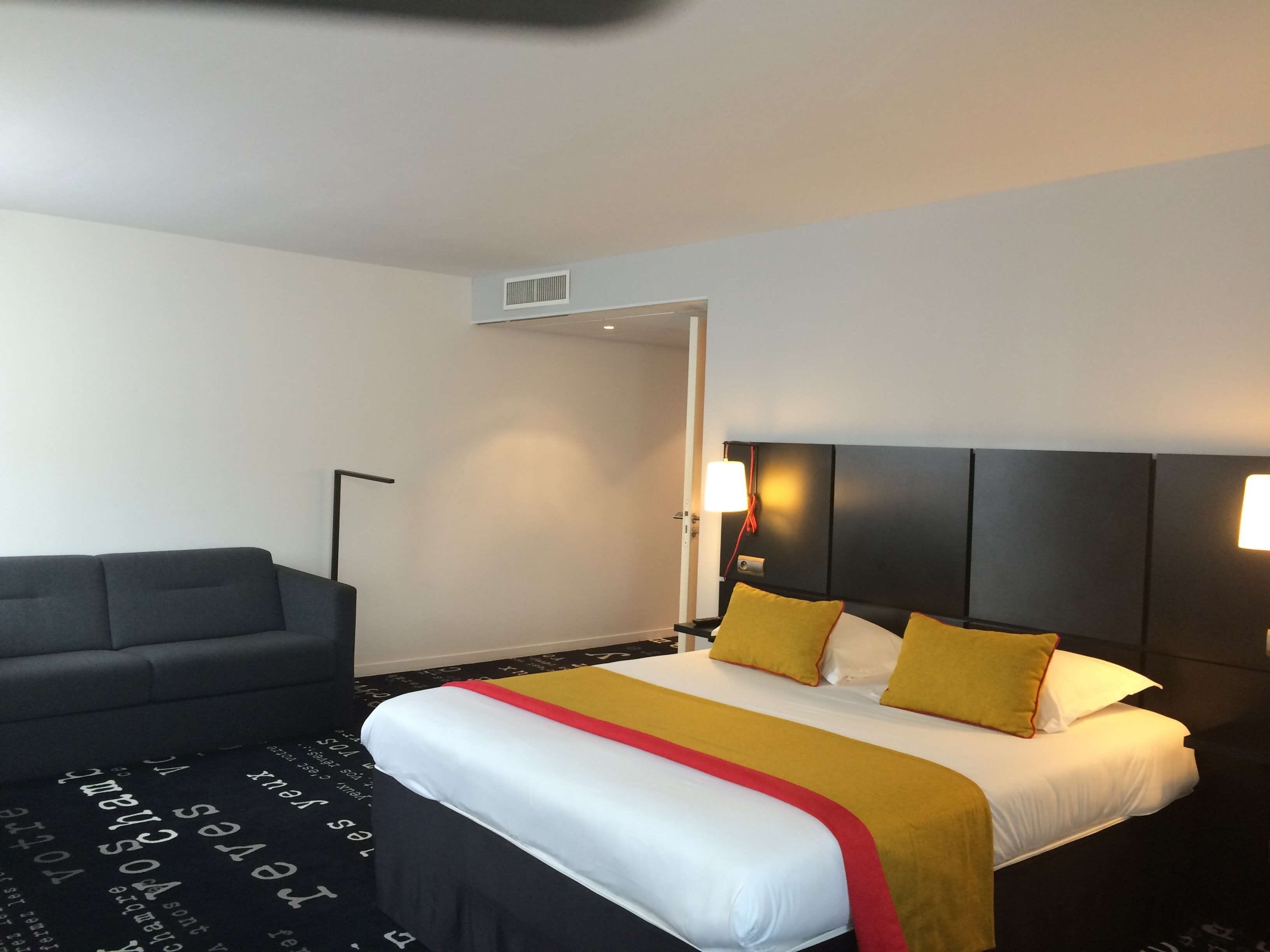 Quarto de hotel com cama, sofá, luminária de chão e um respiradouro na parede.