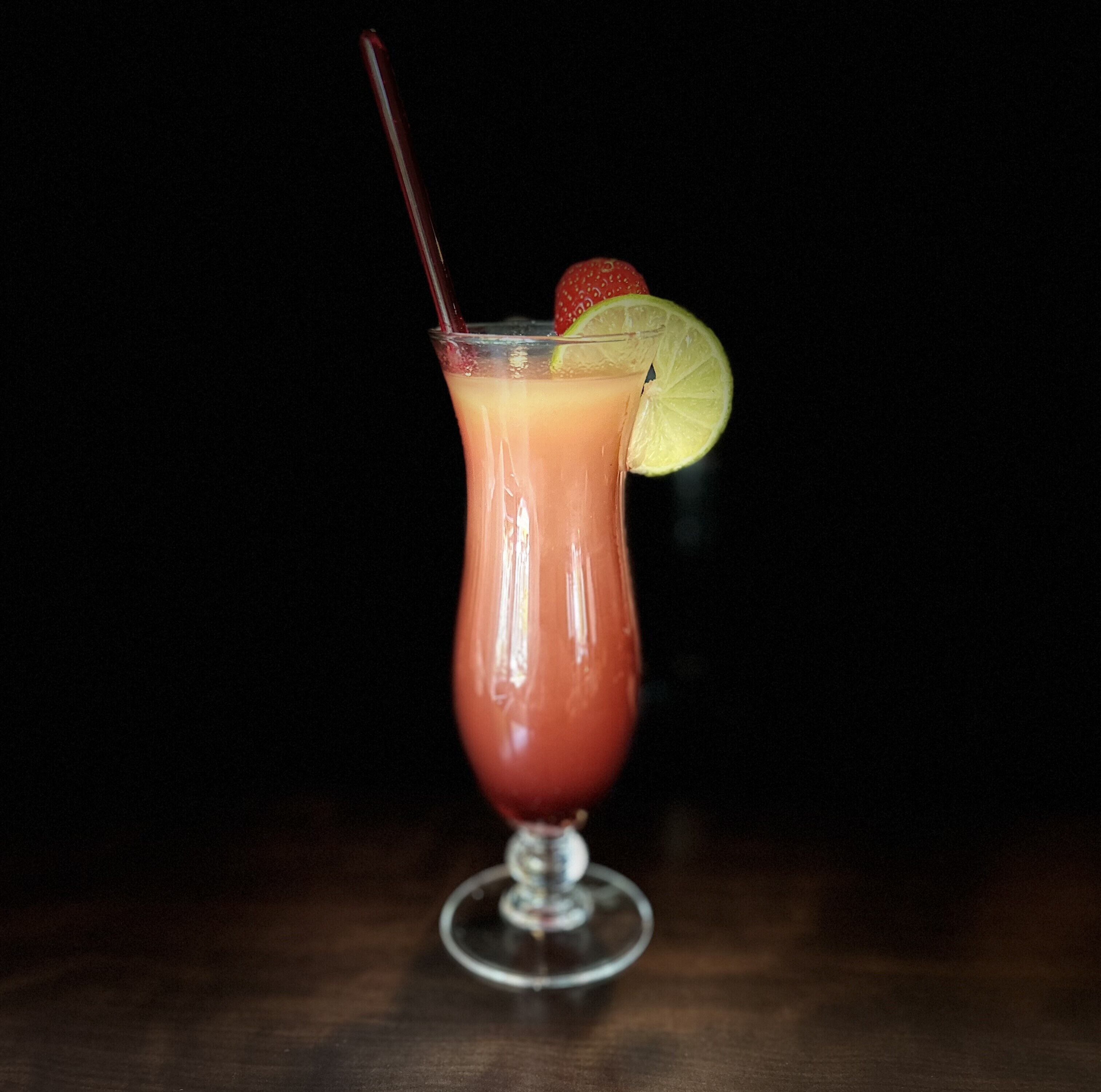 A tall glass with a red drink, garnished with a strawberry and a lime slice, against a dark background.