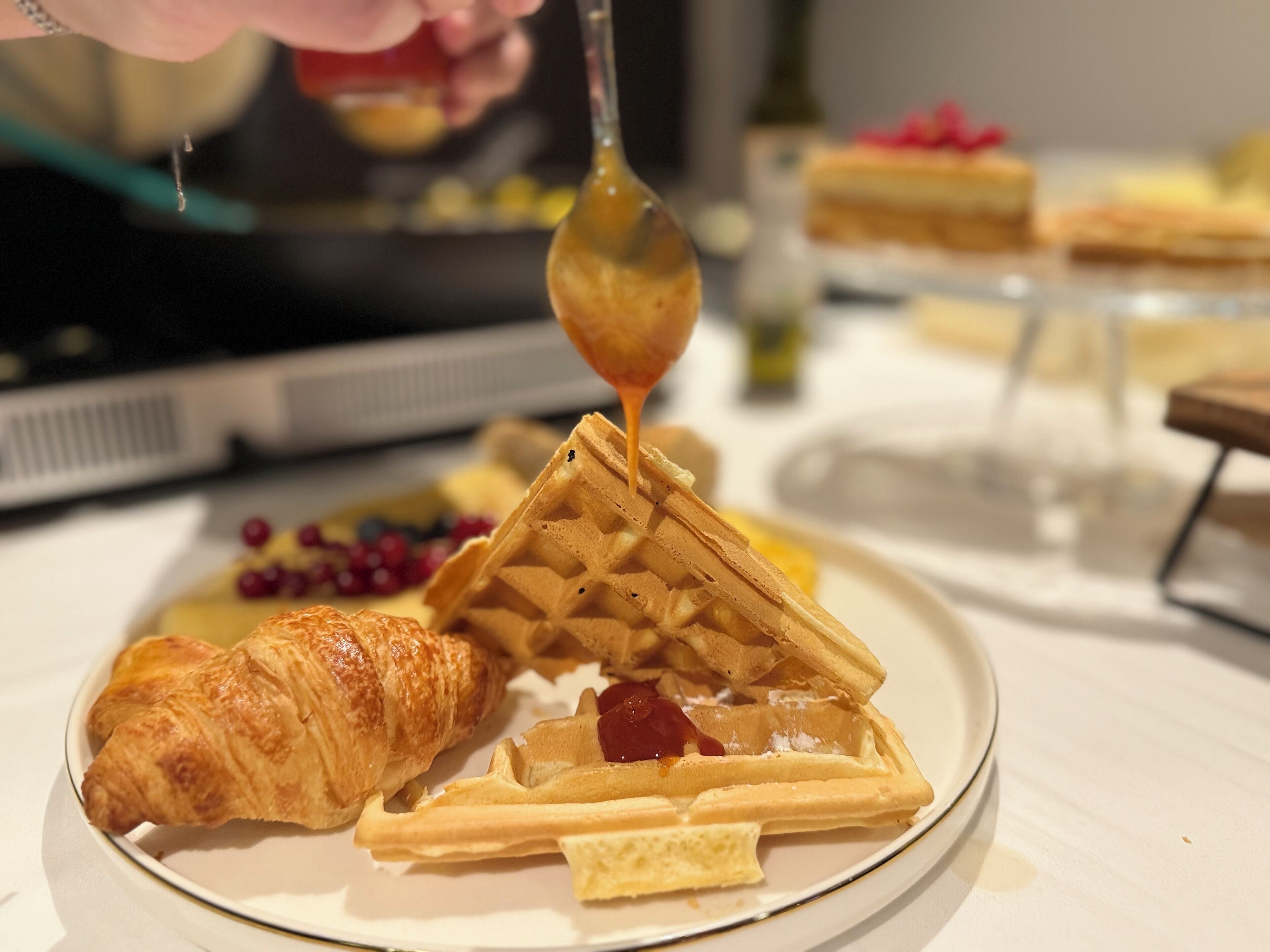 A plate with waffles, a croissant, and berries, with syrup being poured on the waffles.