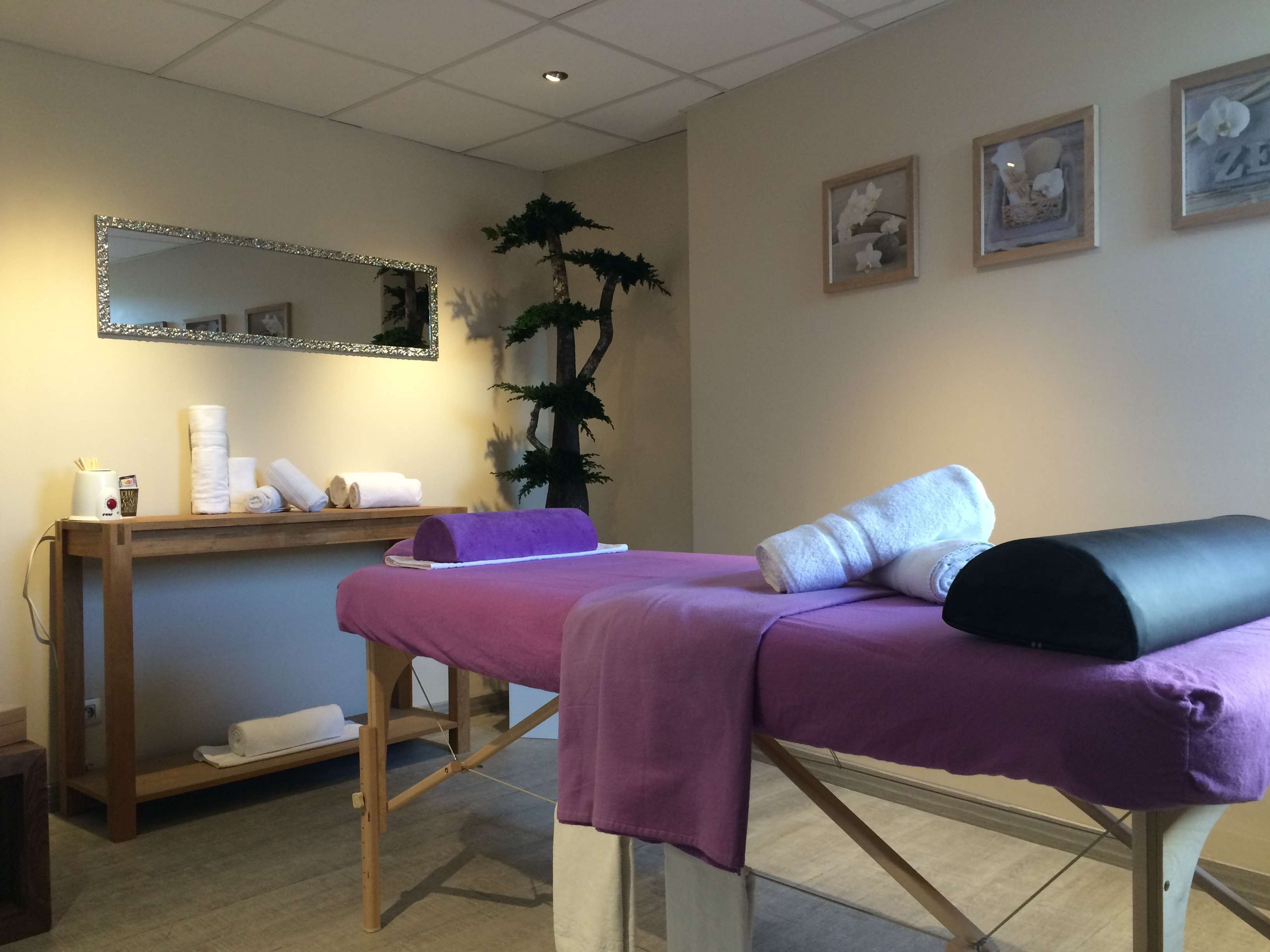 Une table de massage avec une serviette violette et une serviette noire enroulée, un banc en bois avec des serviettes et une tasse, un miroir avec un cadre argenté, et deux tableaux encadrés accrochés au mur.
