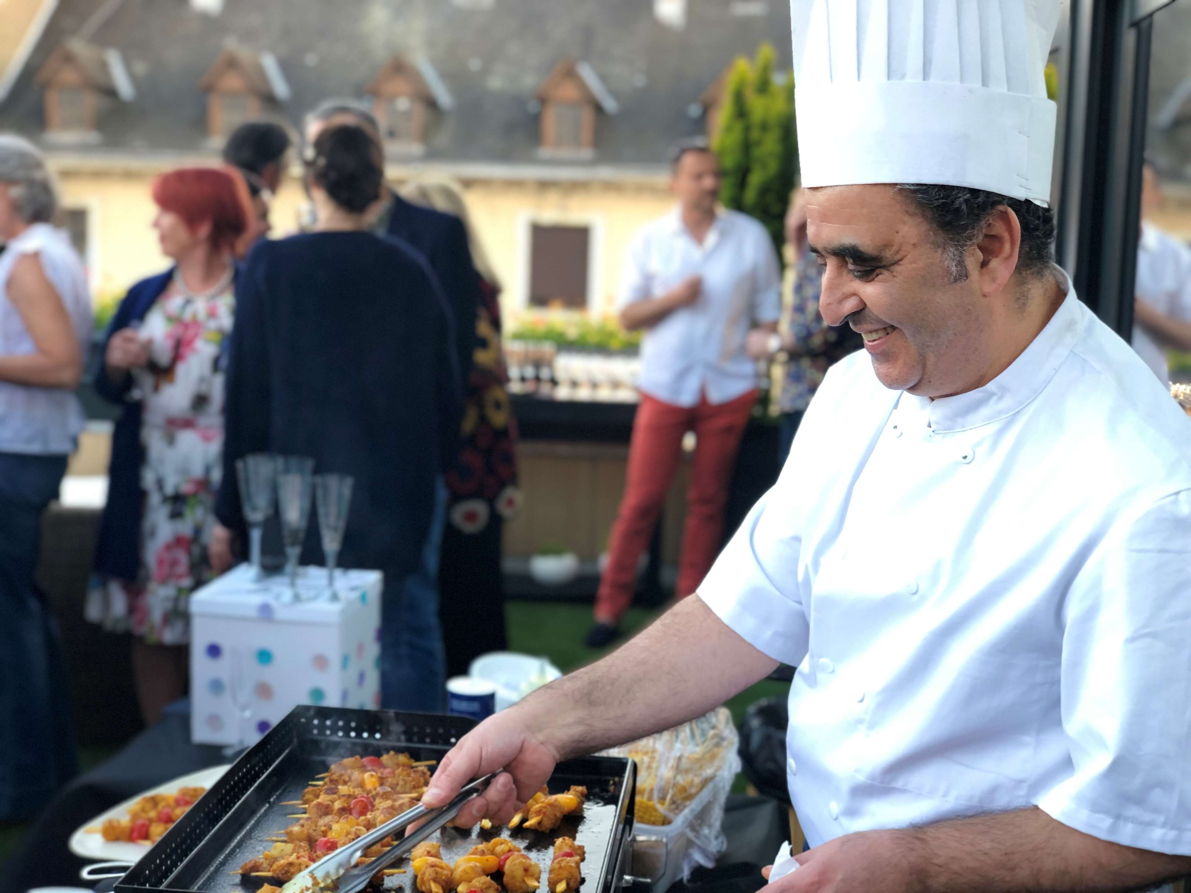 Un chef prepara comida al aire libre con una parrilla, rodeado de gente.