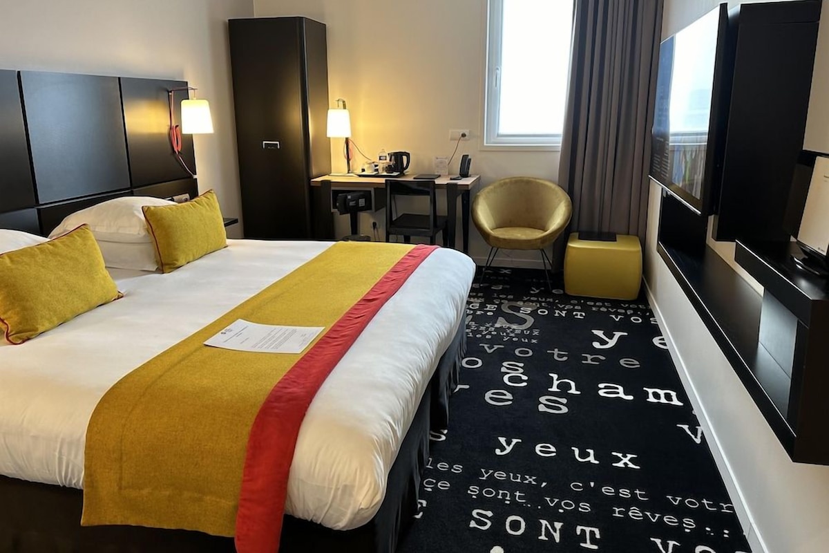 Ein Hotelzimmer mit Bett, Schreibtisch, Sessel, Fernseher und einem Fenster mit Vorhängen.