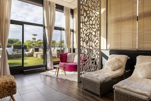Un espacio lounge moderno con un gran ventanal, sillones de mimbre, un sofá rosa y una pantalla decorativa.