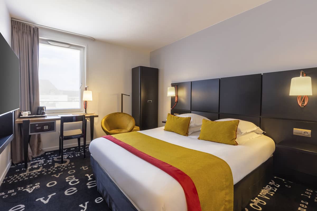 Habitación de hotel con una cama grande, un escritorio con silla, televisión y ventana con cortinas.