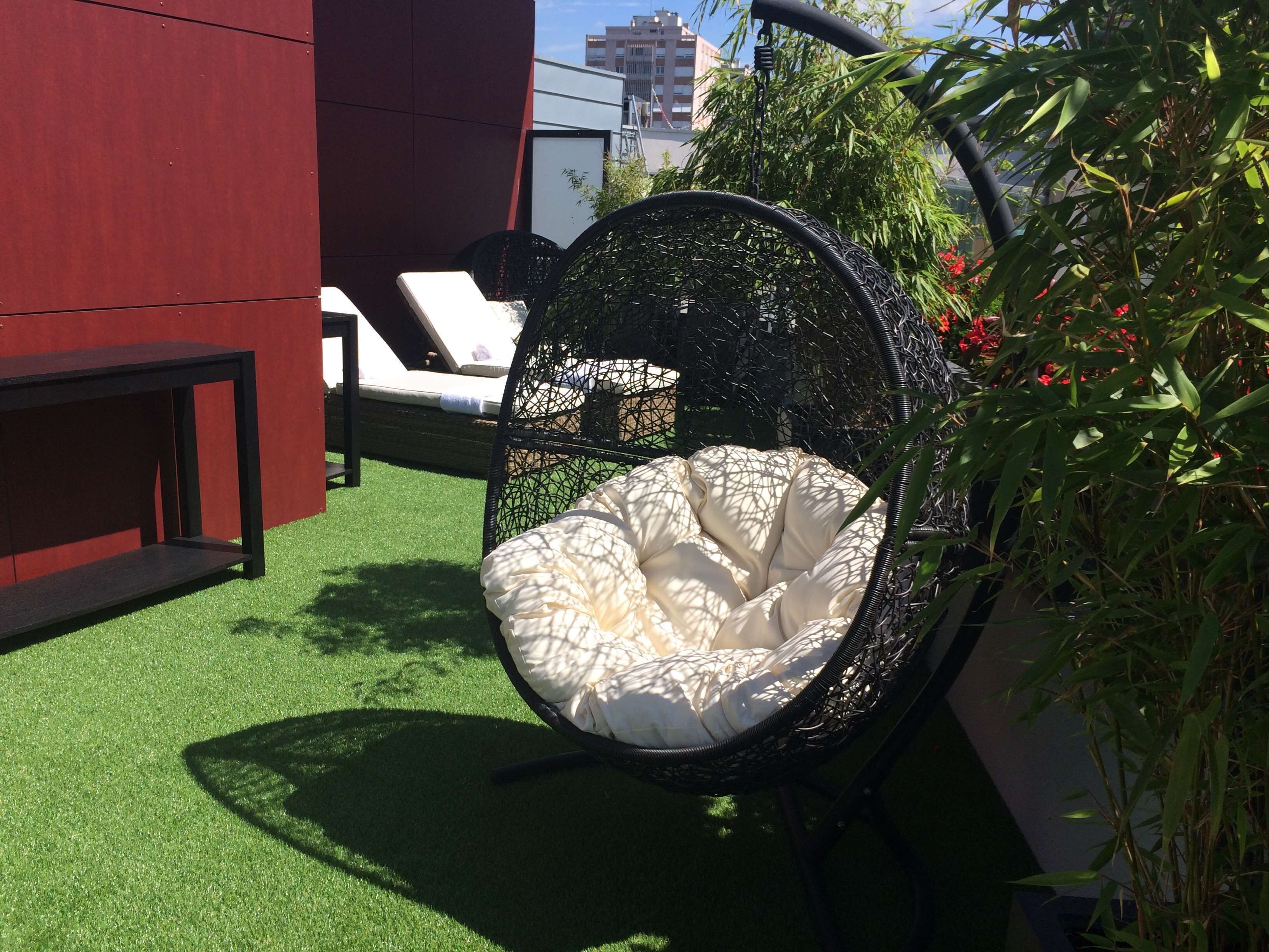 A hanging chair with a cushioned seat and back, positioned on artificial grass, with a red wall and a white chair in the background.