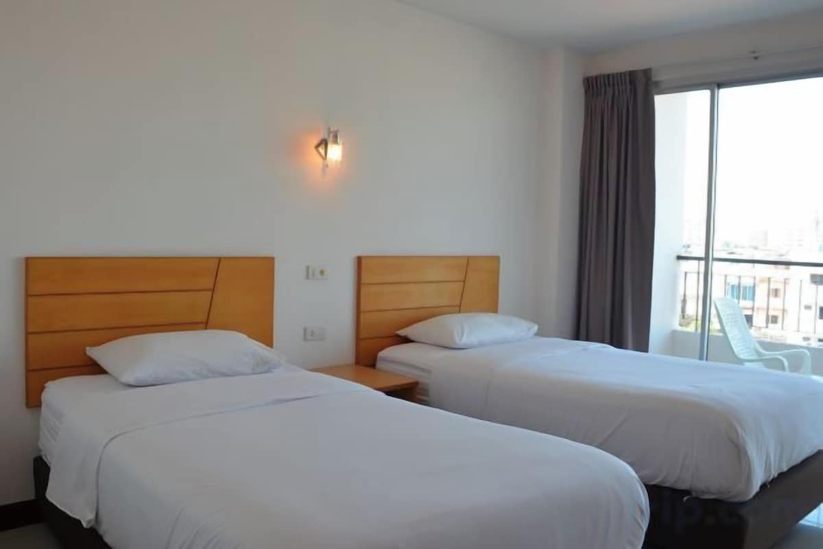 Quarto de hotel com duas camas, mesa de cabeceira, luminária fixada na parede e vista da cidade através de grandes janelas.